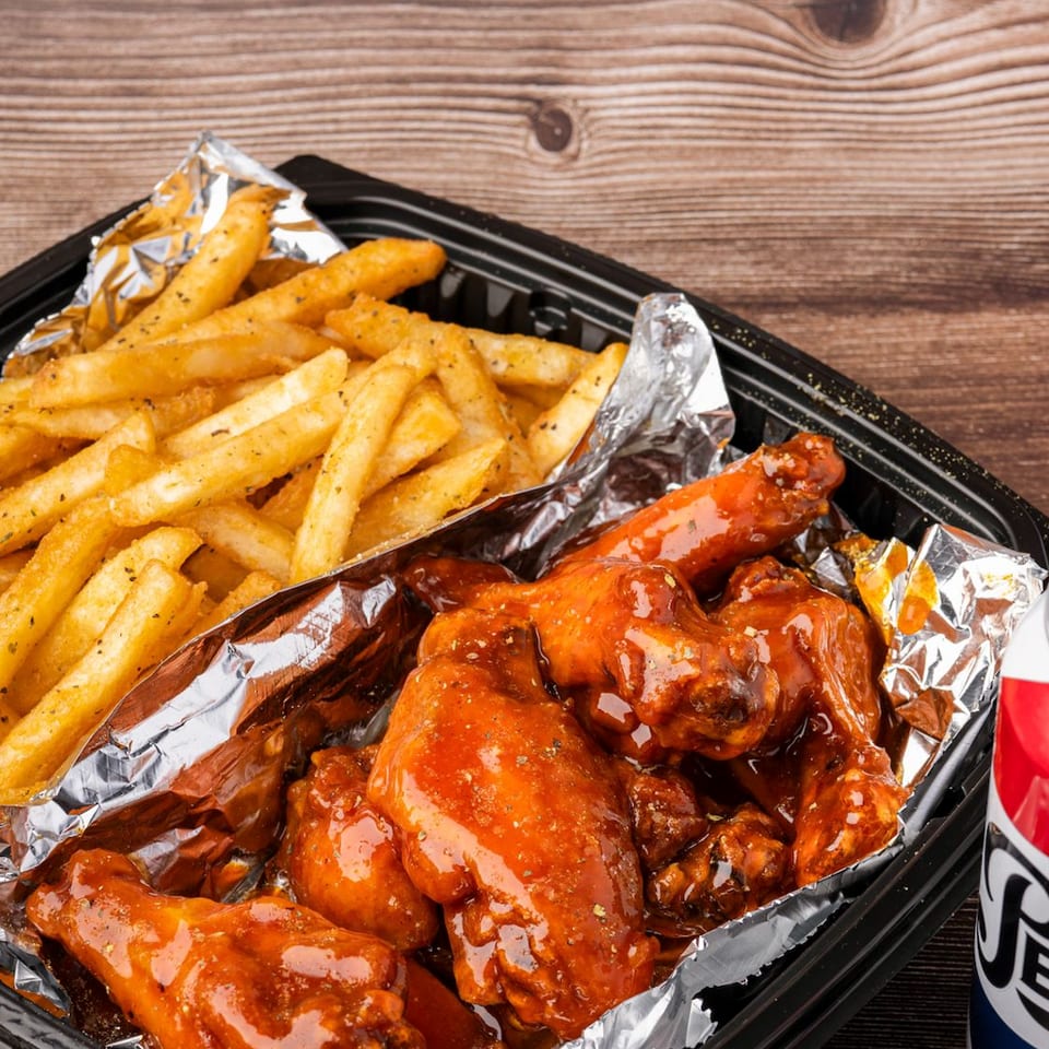 Takeout container with golden french fries, saucy chicken wings, and a Pepsi can on a wooden table.