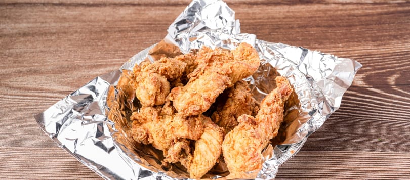 Crispy fried chicken tenders in a foil-lined container on a wooden table.