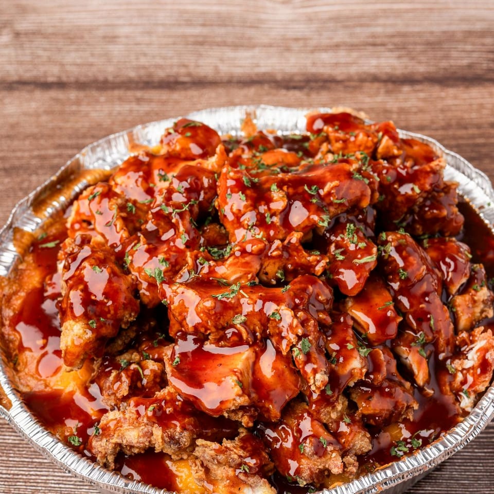 Crispy chicken bites drenched in a rich, dark sauce and sprinkled with herbs, in a foil container.