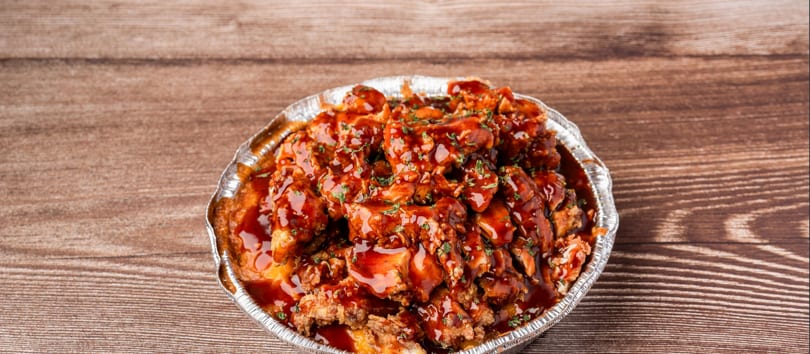 Crispy chicken bites drenched in a rich, dark sauce and sprinkled with herbs, in a foil container.