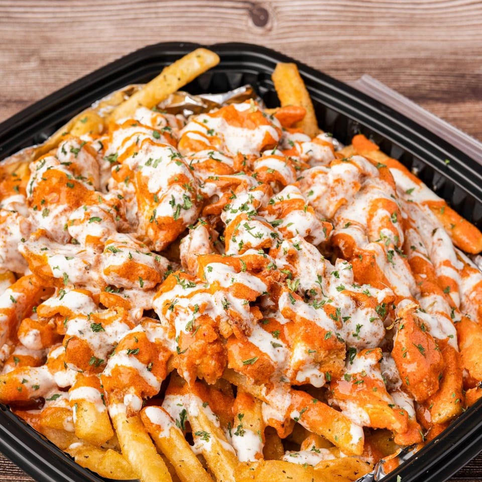 A black takeout container filled with golden french fries, topped with orange sauce, white drizzle, and green herbs.