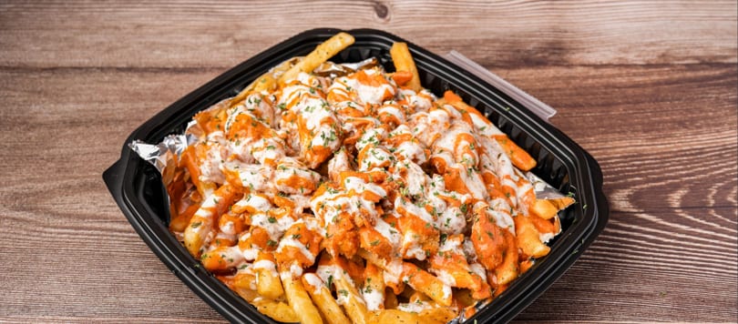 A black takeout container filled with golden french fries, topped with orange sauce, white drizzle, and green herbs.