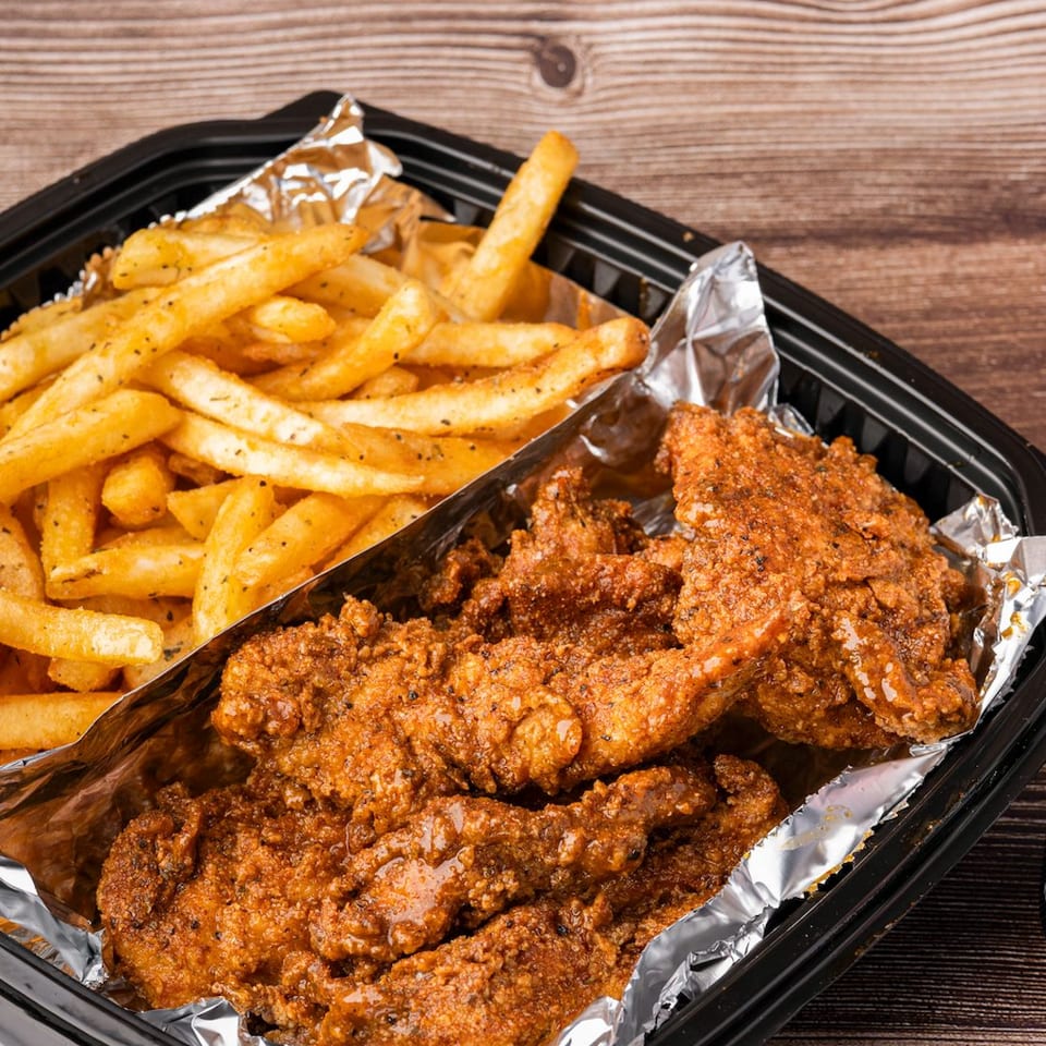 Crispy fried chicken tenders and seasoned fries in a black takeout container with a Pepsi-Cola can.